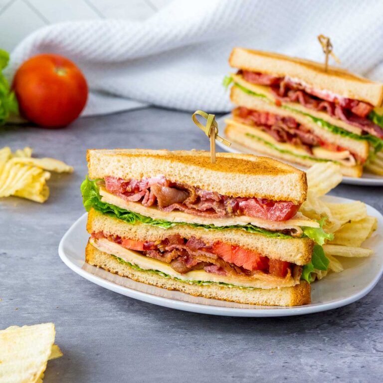 Chicken club sandwich cut into two and served with chips on white plates.