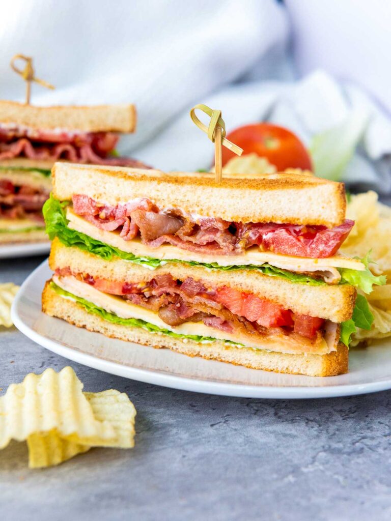 Chicken club sandwich cut into two and served with chips on white plates. There are some chips on the grey surface in front of the plate.