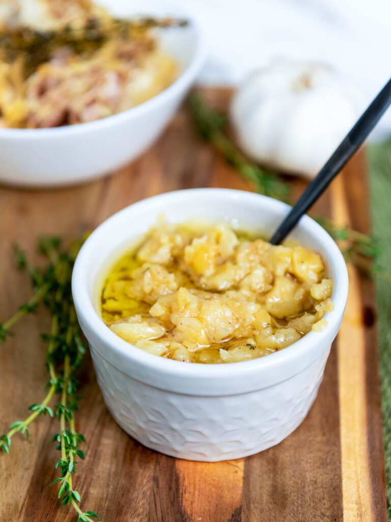 A bowl of garlic confit on a wooden board with roasted garlic peel and thyme leaves in the background.
