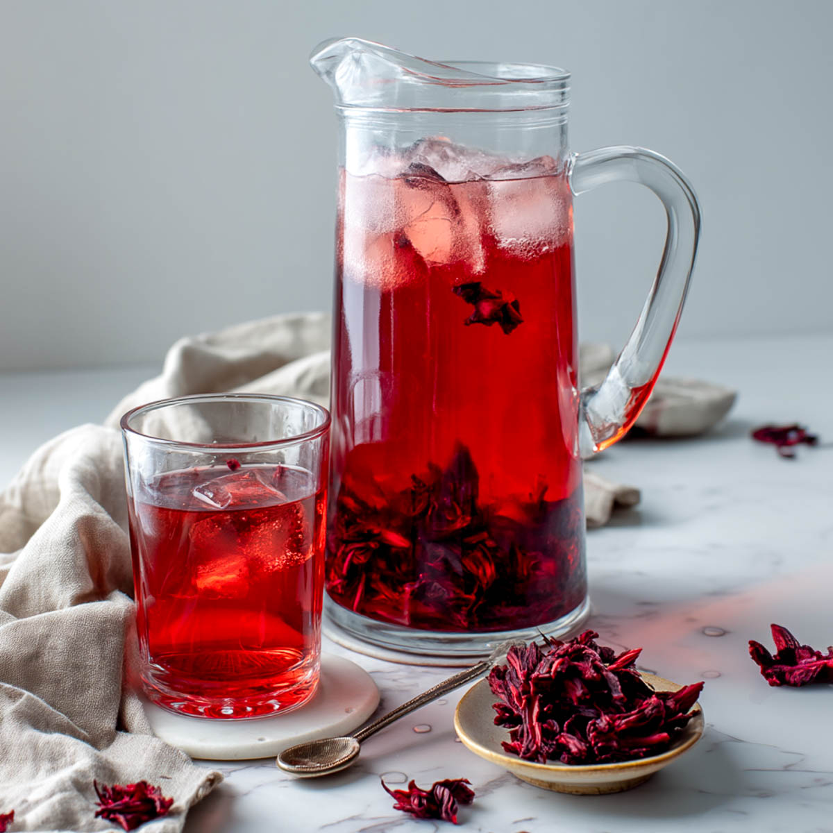 Pitcher of cold brew hibiscus water with ice and strained pink infusion, beside dried petals and a mauve napkin on white marble.