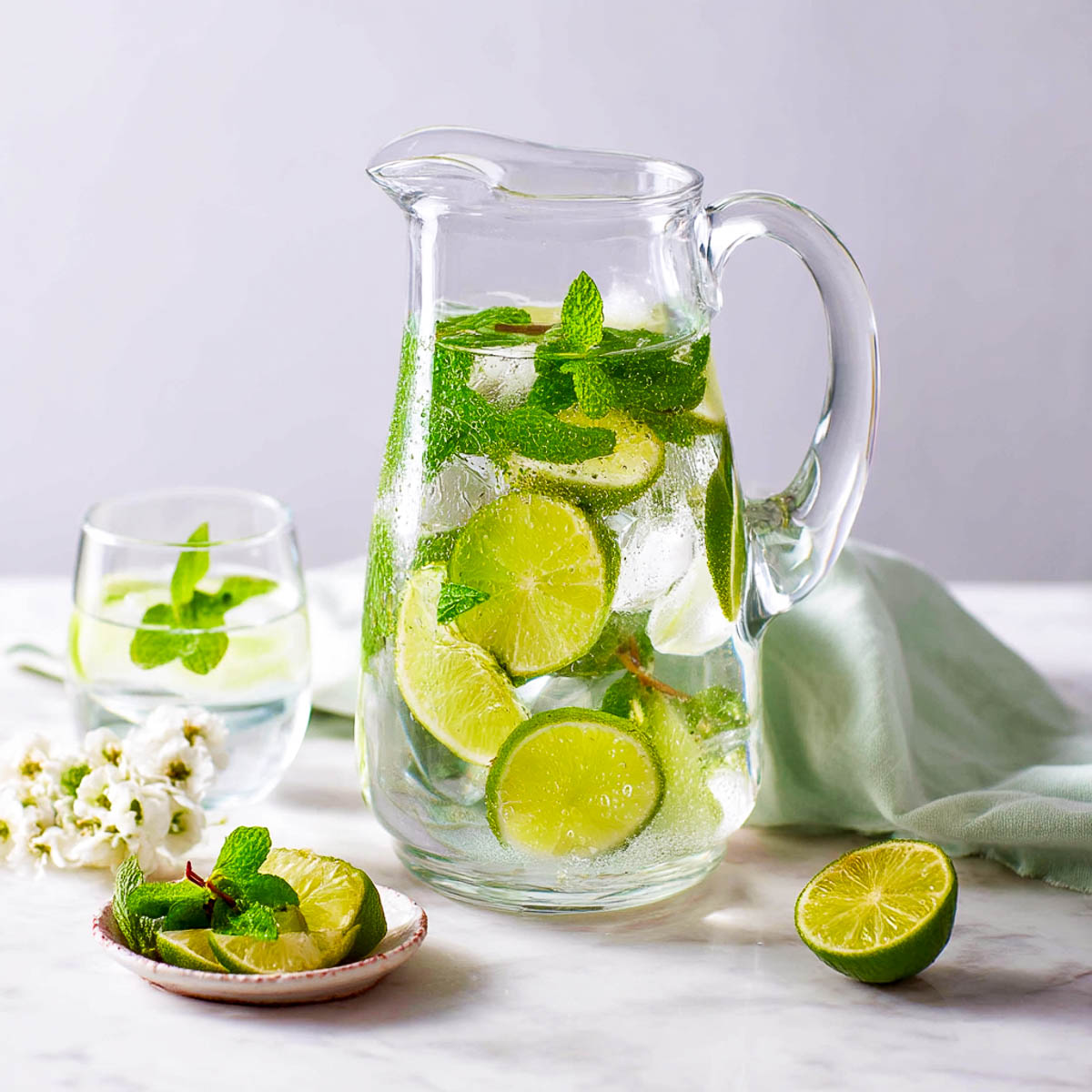 Pitcher and glass of lime mint water with ice, soft mint napkin, lime slices, and fresh mint on a white marble surface.