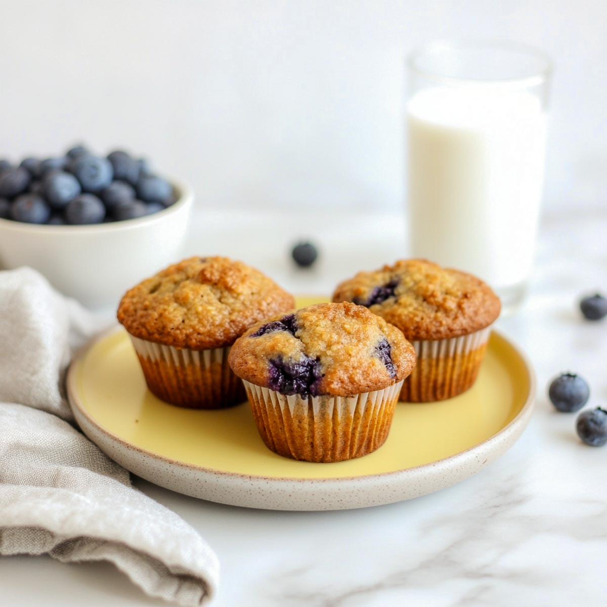 Three golden low-sugar blueberry muffins on yellow plate with blueberries.