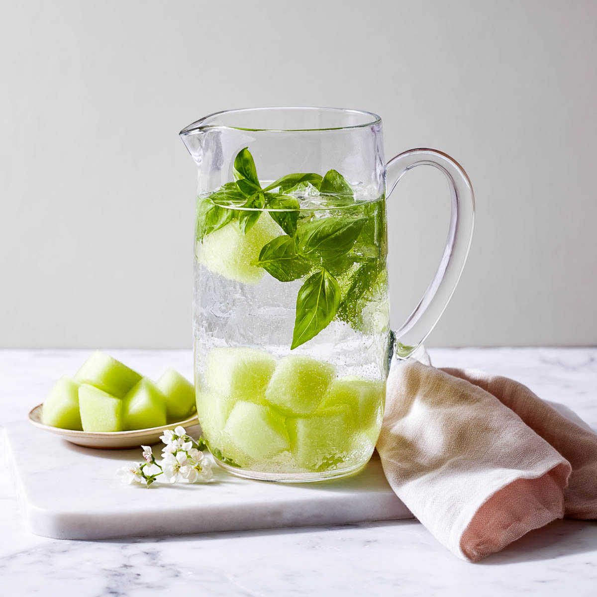 Pitcher of melon basil infused water with pale green melon cubes, basil leaves, and a soft linen napkin on marble background.