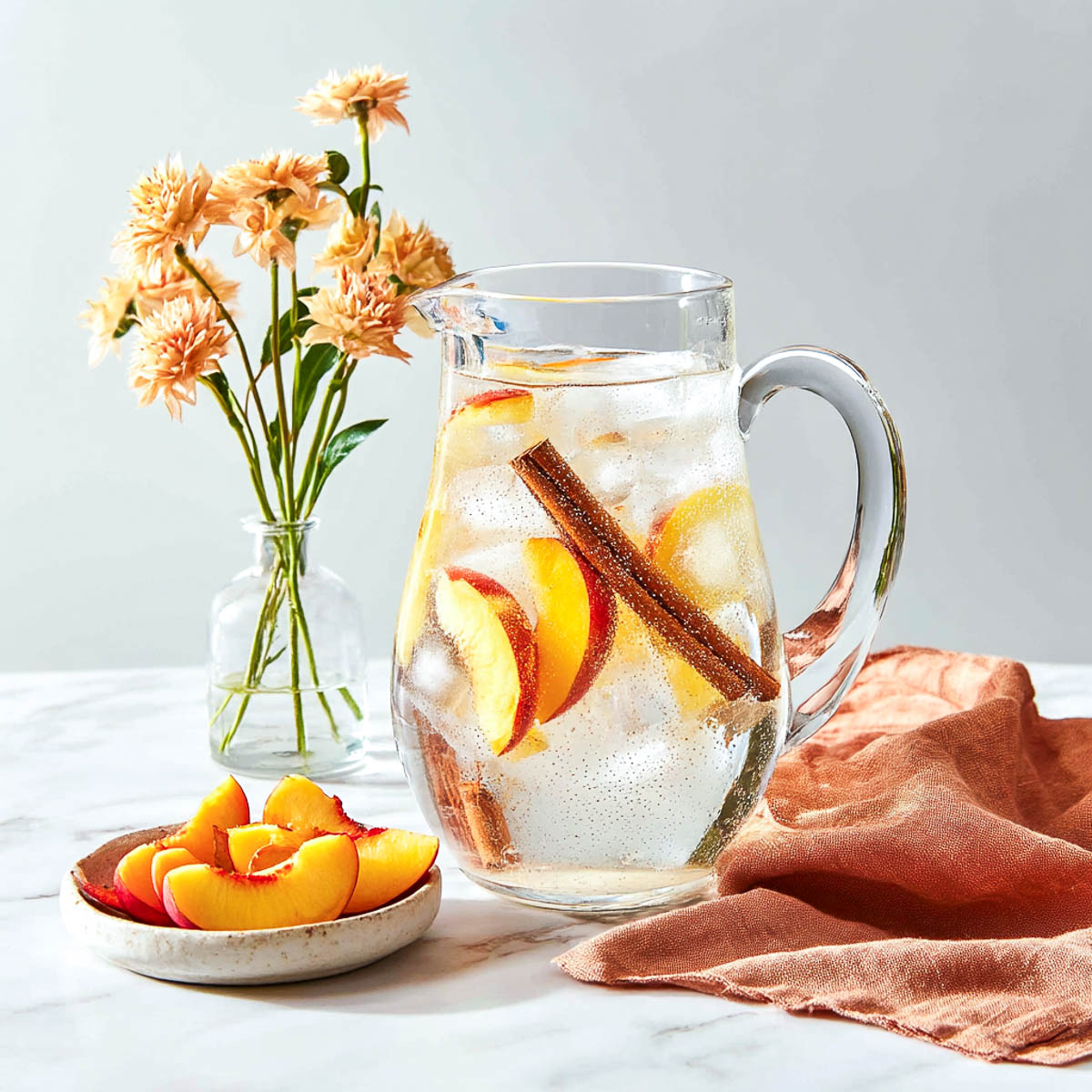 Pitcher of peach cinnamon water with sliced peaches, submerged cinnamon stick on marble background beside a small vase of warm-toned flowers.