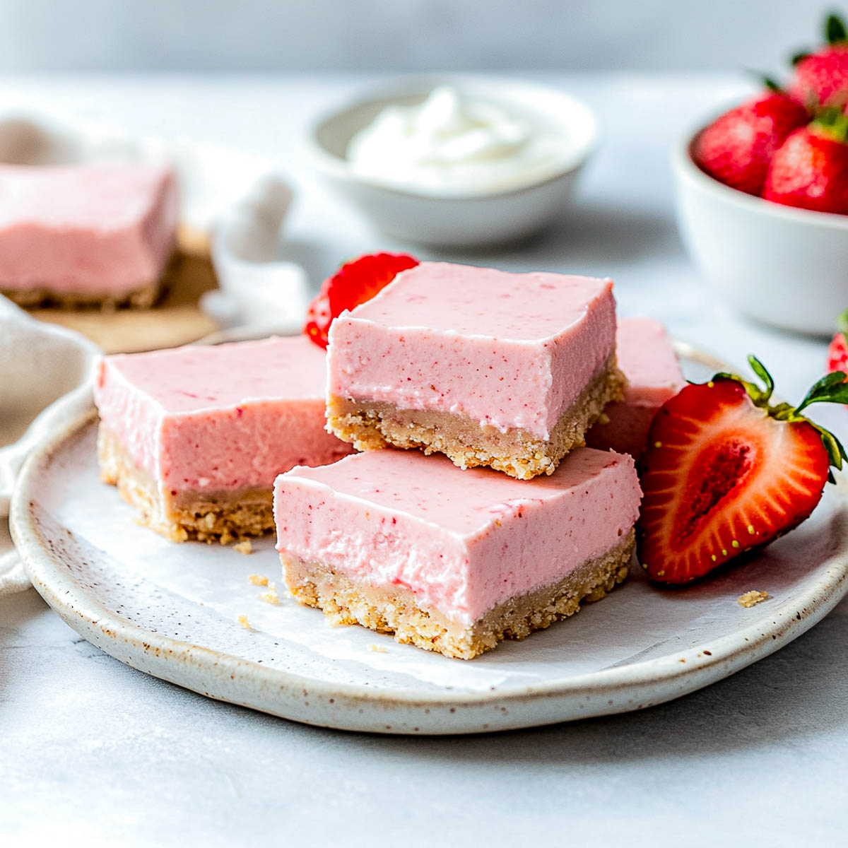 Strawberry cheesecake bars on a white plate with a bowl of fresh strawberries in the background.