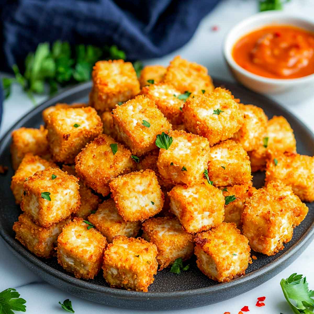Crispy tofu nuggets stacked on a dark plate with dipping sauce on the side.