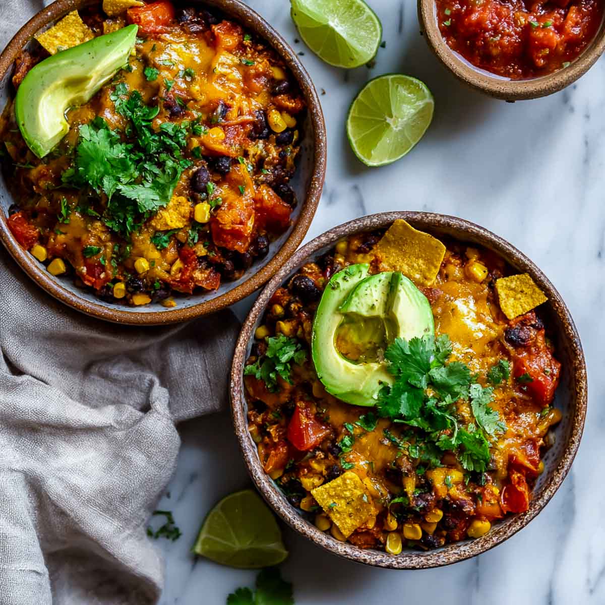 Top-down view of two bowls with cheesy enchilada filling, avocado slices, and lime wedges.