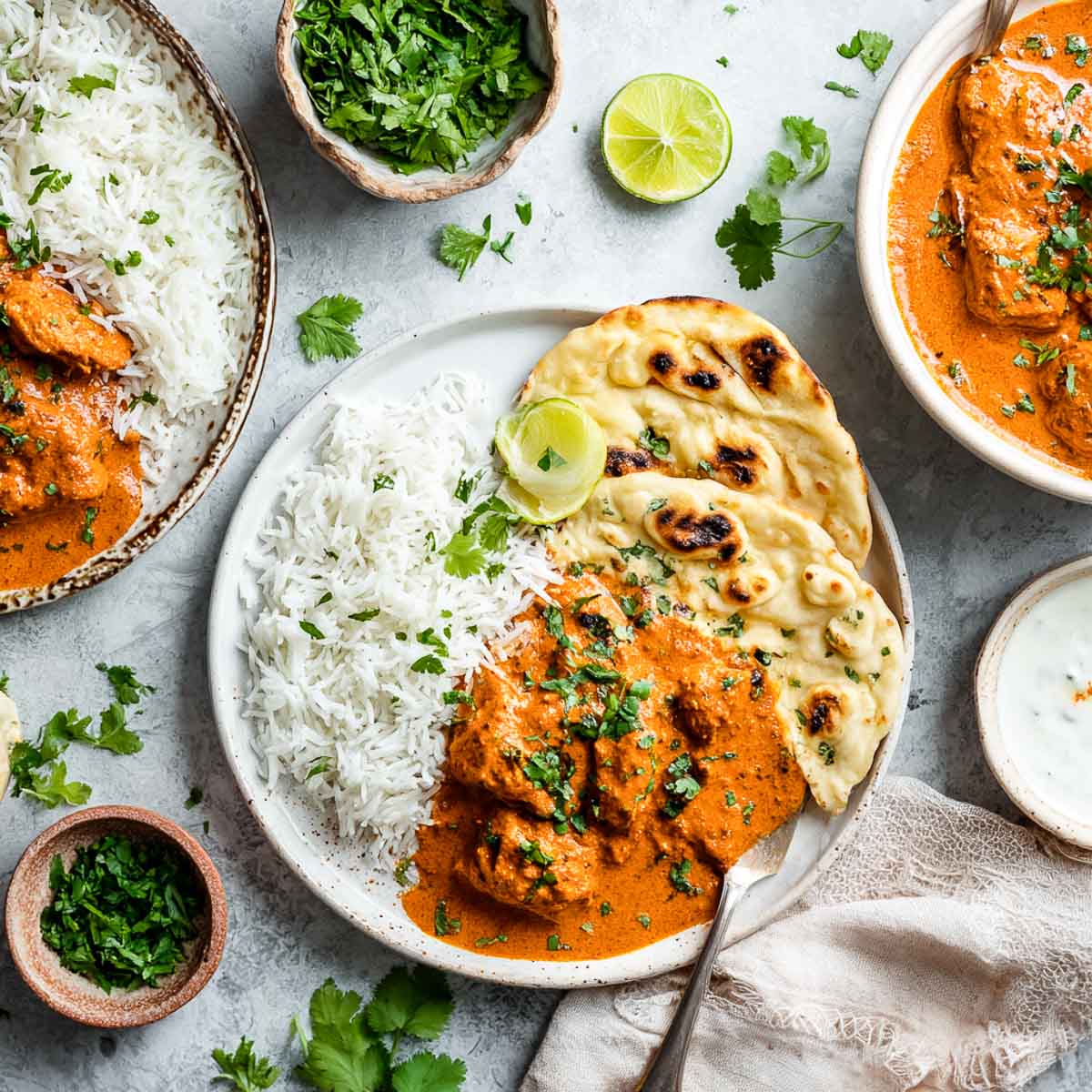 Butter chicken served with rice and naan on a white plate, with fresh cilantro and butter chicken in a serving bowl in the background.