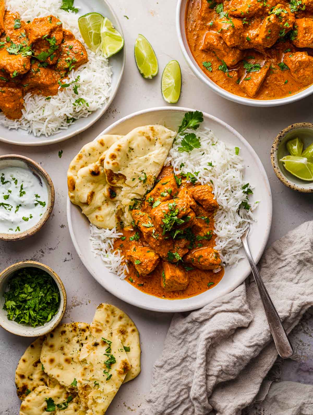 Overhead view of a plate of butter chicken with basmati rice and naan, surrounded by lime wedges, cilantro, and yogurt sauce.