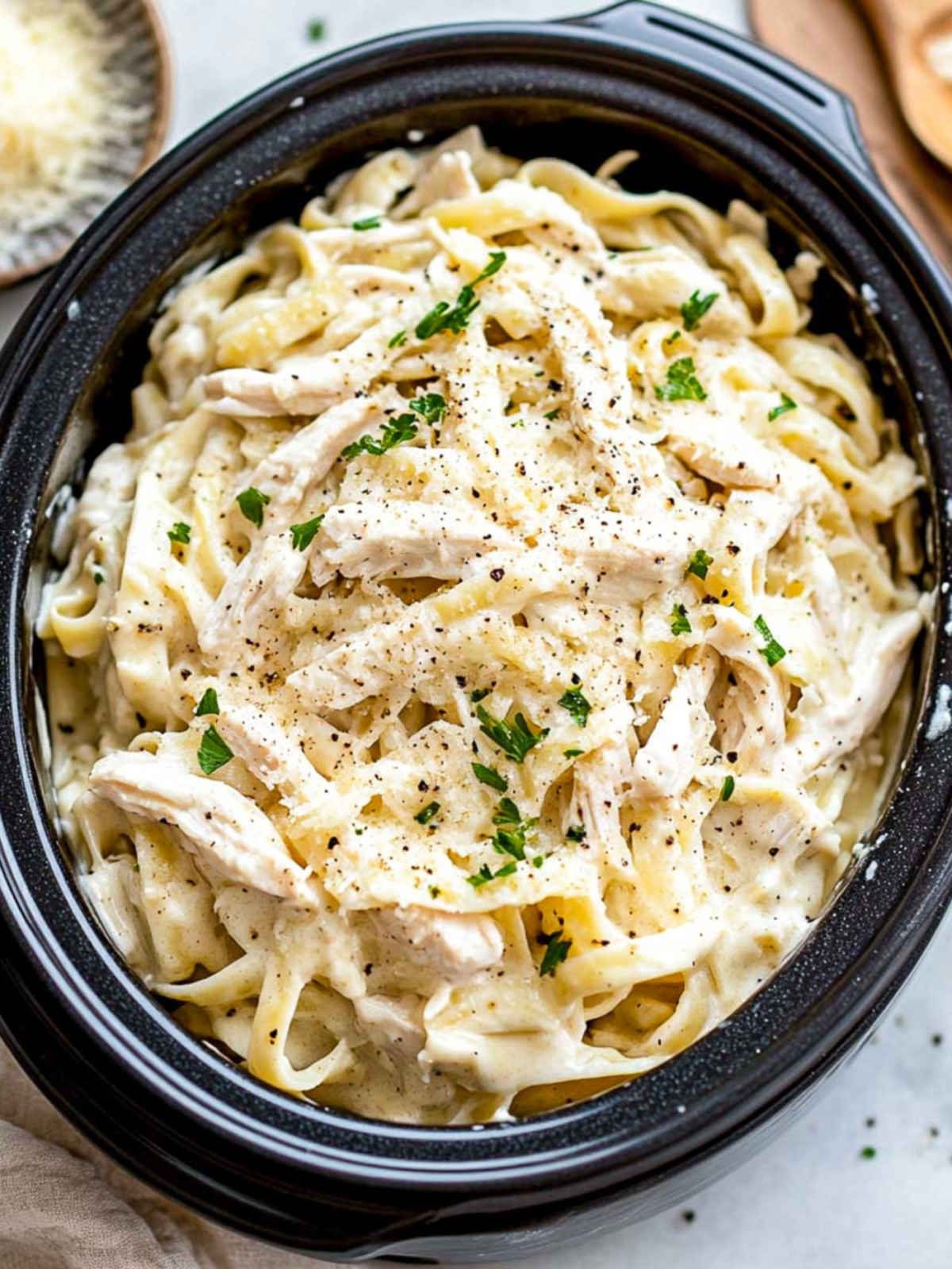Slow cooked chicken Alfredo in a black crockpot, topped with parmesan and parsley.
