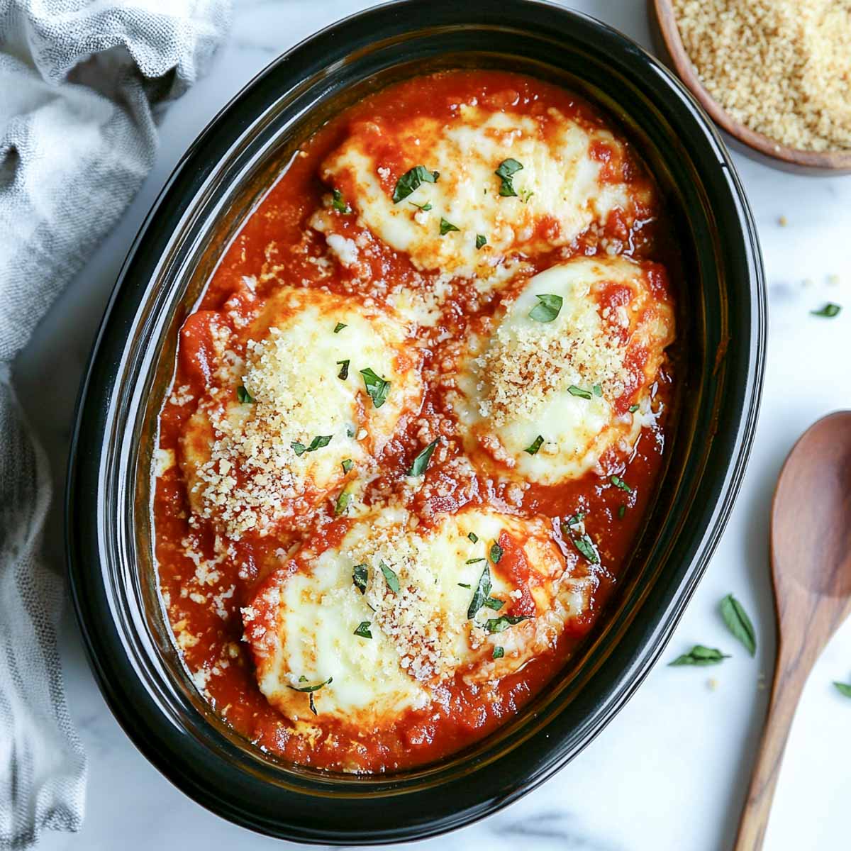 Crockpot Chicken Parmesan in a black slow cooker topped with melted cheese, marinara sauce, and fresh basil.