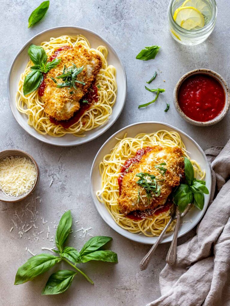 Top view of Chicken Parmesan with spaghetti on two plates, surrounded by fresh basil, shredded cheese, and a napkin.