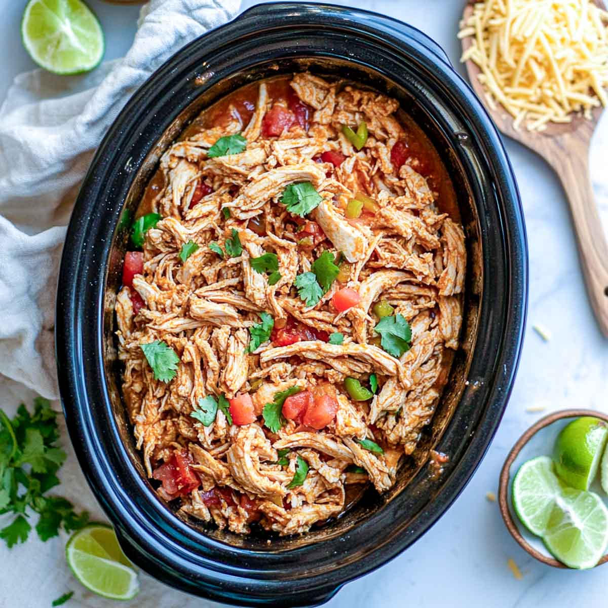 Shredded seasoned chicken in a black Crockpot with tomatoes and fresh cilantro.