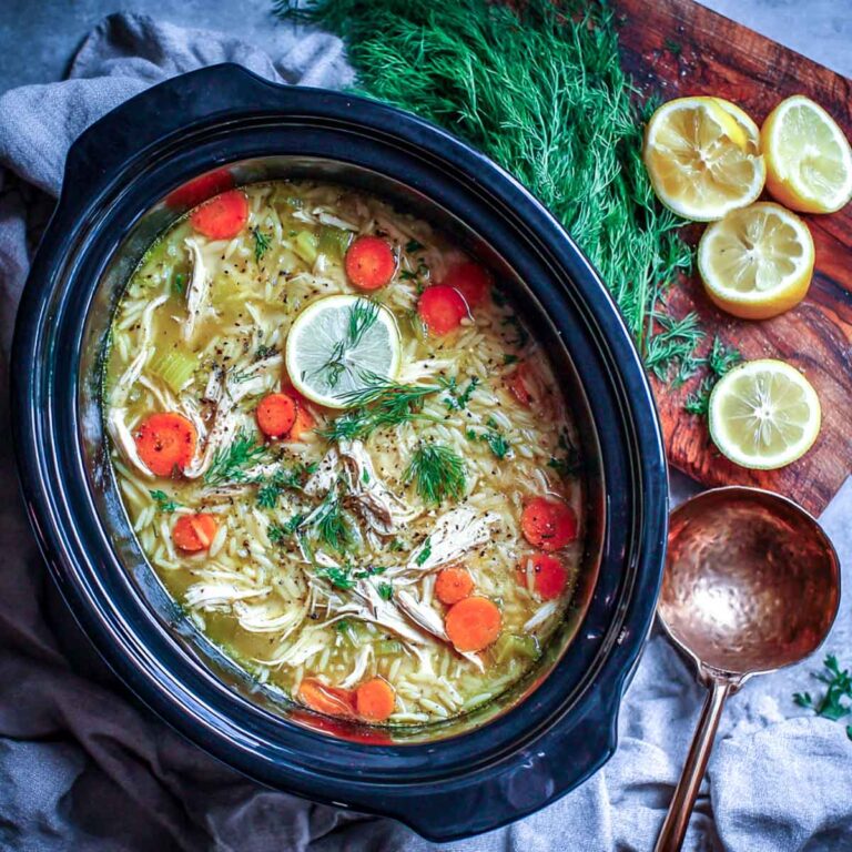 Crockpot filled with lemon chicken orzo soup, surrounded by fresh dill, lemon slices, and a copper ladle on a rustic cutting board.