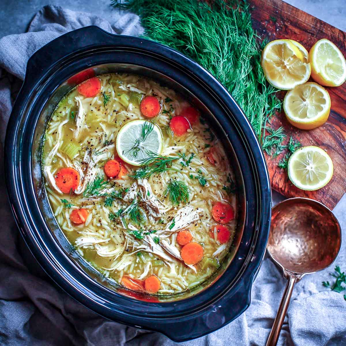 Crockpot filled with lemon chicken orzo soup, surrounded by fresh dill, lemon slices, and a copper ladle on a rustic cutting board.