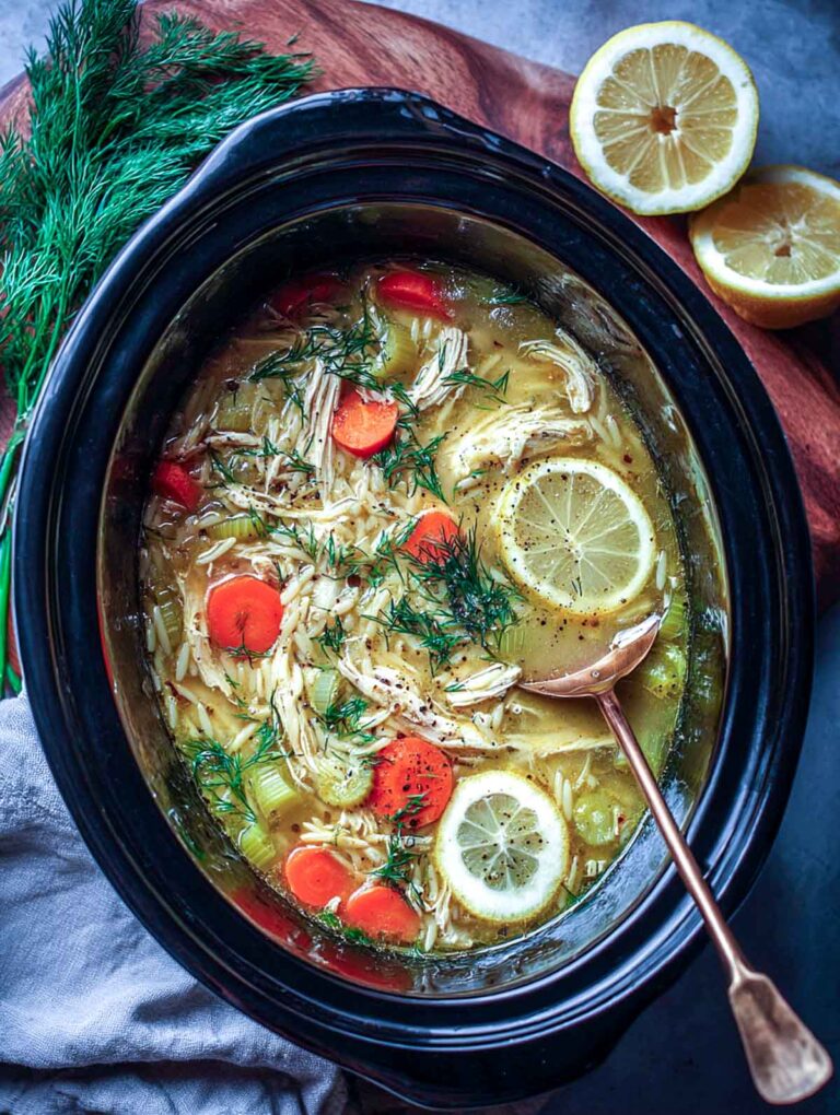 Top view of crockpot lemon chicken orzo soup with sliced lemons, dill, and a vintage soup ladle resting in the pot.