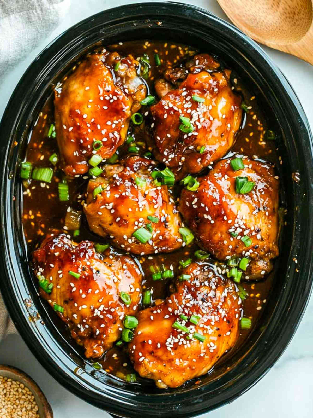 Slow-cooked chicken thighs in a sticky honey garlic sauce, topped with scallions and sesame seeds in a crockpot.