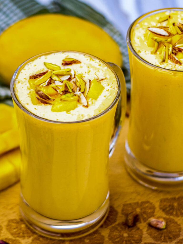 Tall glass of mango lassi topped with chopped pistachios and mango chunks, placed on a wooden board with a ripe mango in the background.