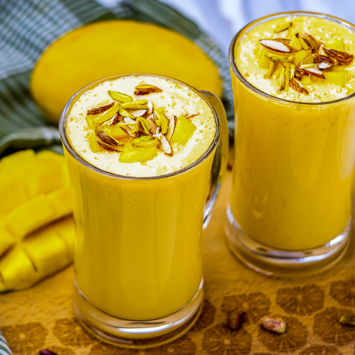 Two glasses of mango lassi garnished with slivered almonds, pistachios, and mango pieces, set against a background of a halved mango and green cloth.