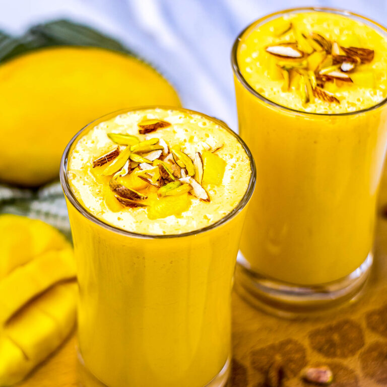 Close-up of mango lassi in glass mugs with creamy tops, garnished with chopped nuts and mango, surrounded by sliced mango and a whole fruit.