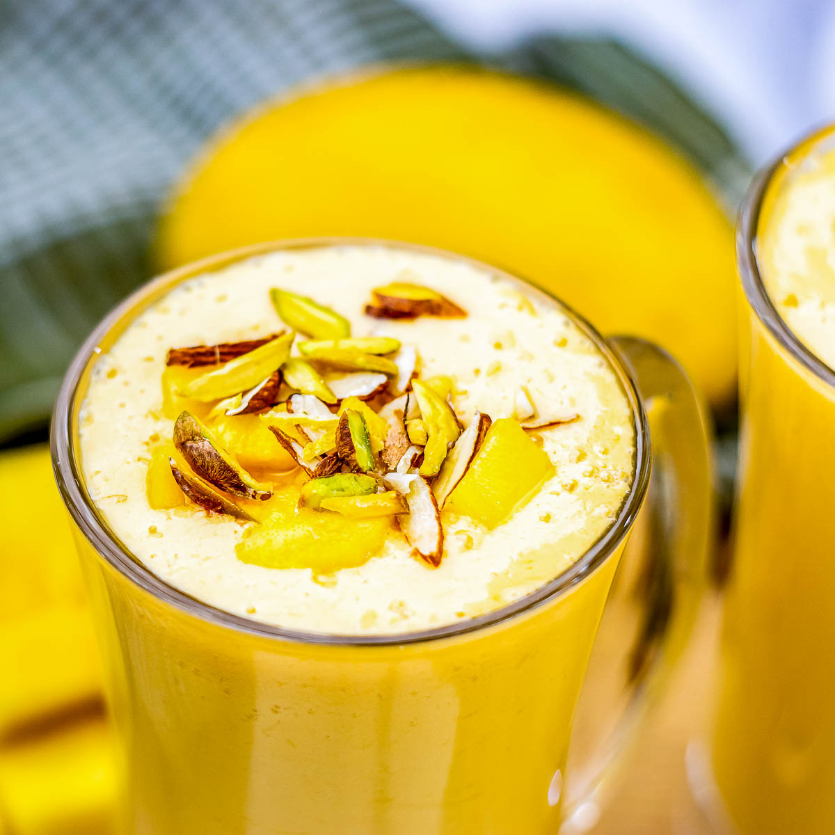 Top view of a mango lassi mug filled to the rim and topped with pistachios, almonds, and fresh mango, with a blurred mango in the background.