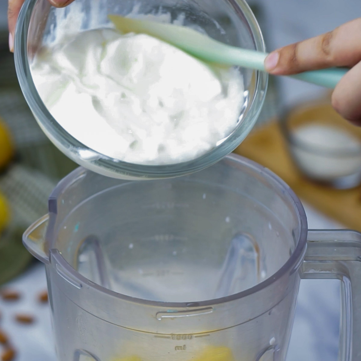 Adding a bowl of yogurt into a blender jar over chopped mangoes, preparing the lassi base.