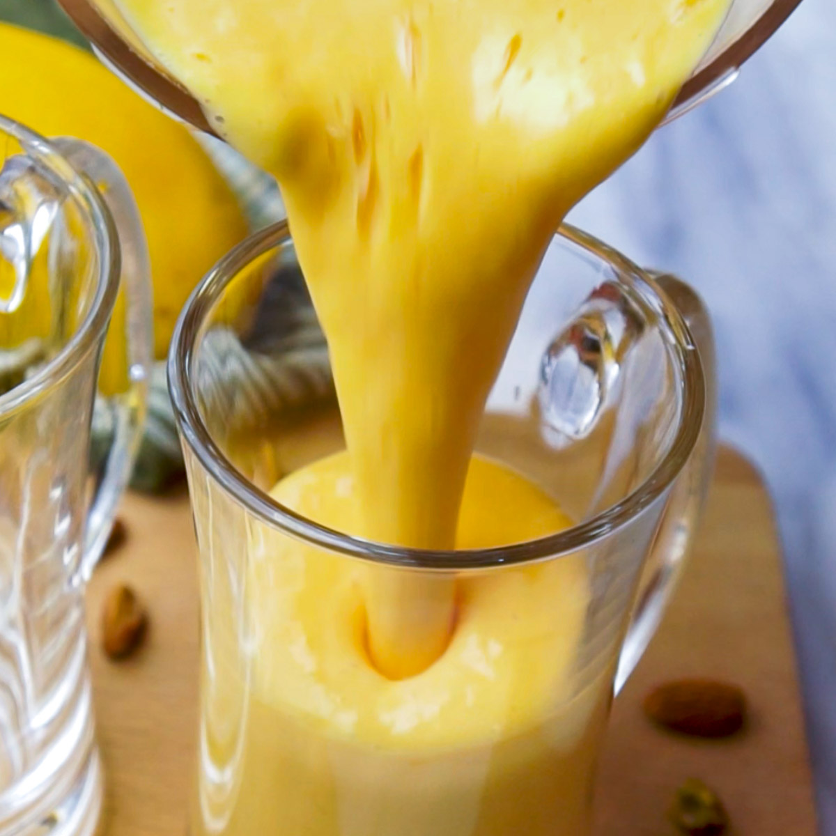 Pouring thick mango lassi from a blender into a clear glass mug, with pistachios scattered on the table.