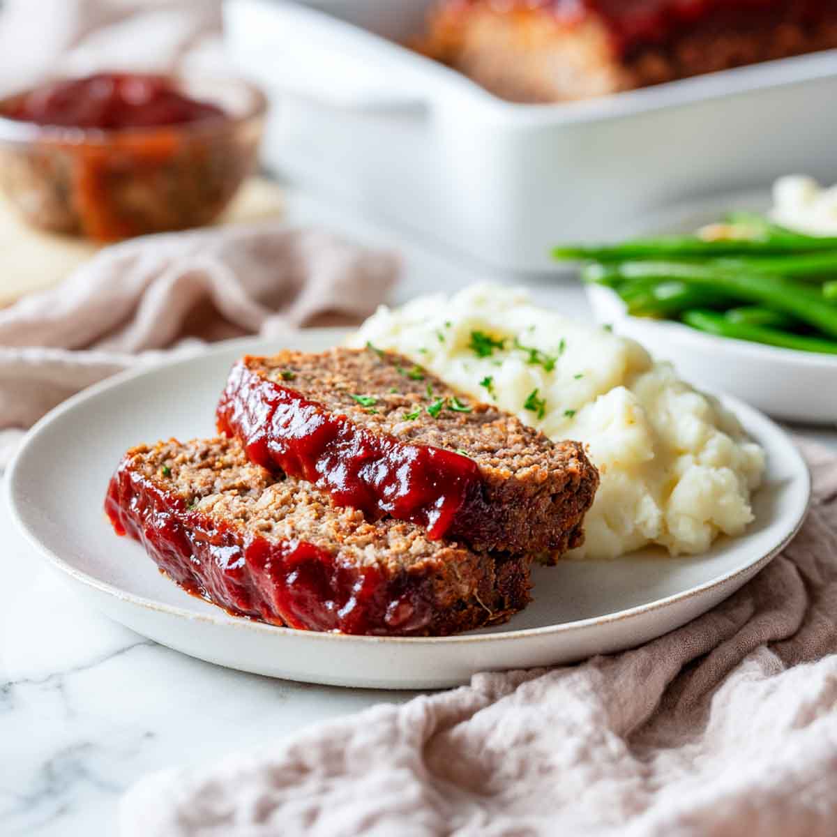 Sliced meatloaf with glossy red glaze served with mashed potatoes and green beans.
