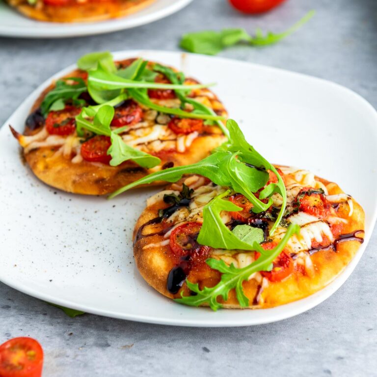Mini Caprese pizzas on a white plate, topped with melted mozzarella, cherry tomatoes, arugula, and a drizzle of balsamic glaze.