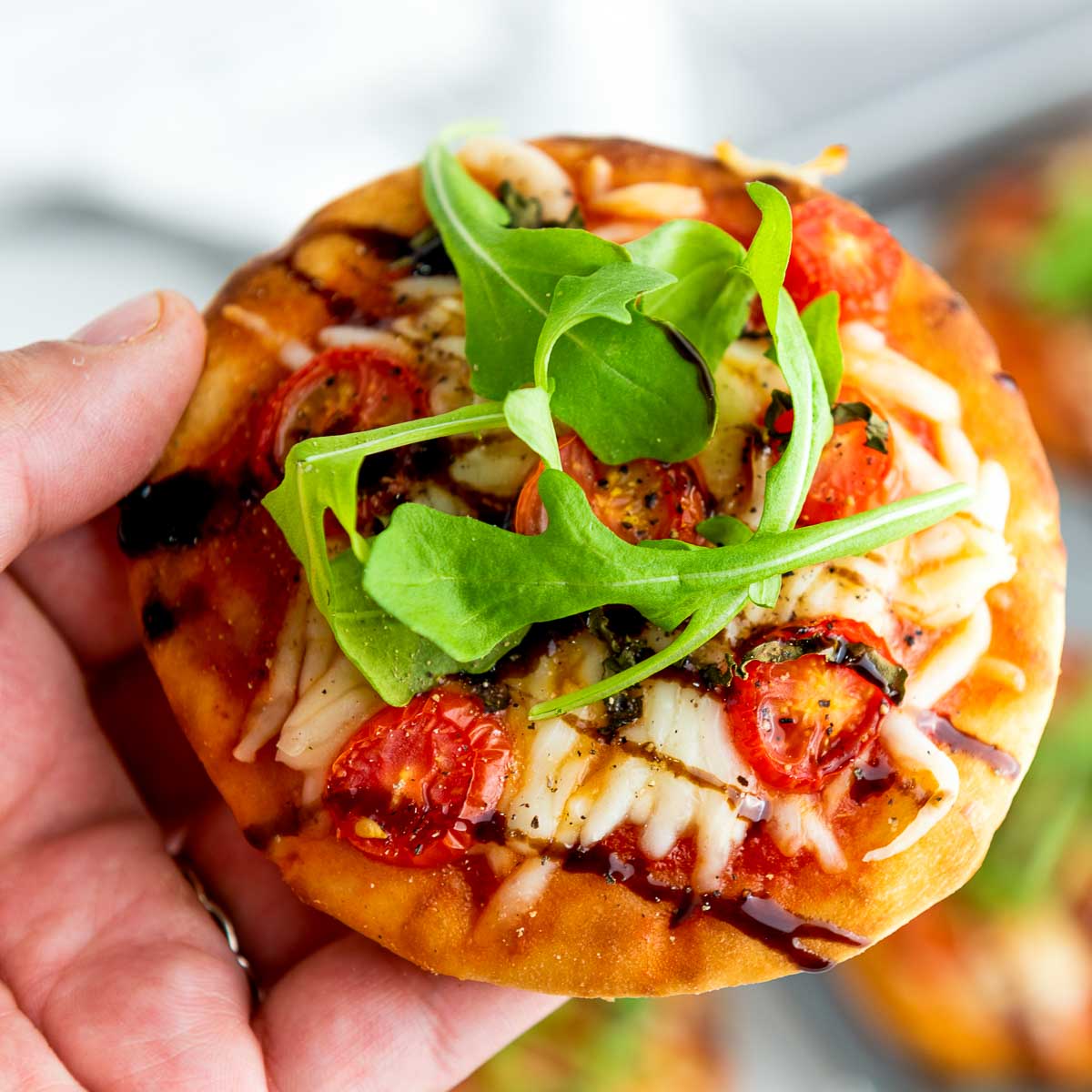Close-up of a hand holding a single mini Caprese pizza topped with arugula, tomatoes, cheese, and balsamic reduction.