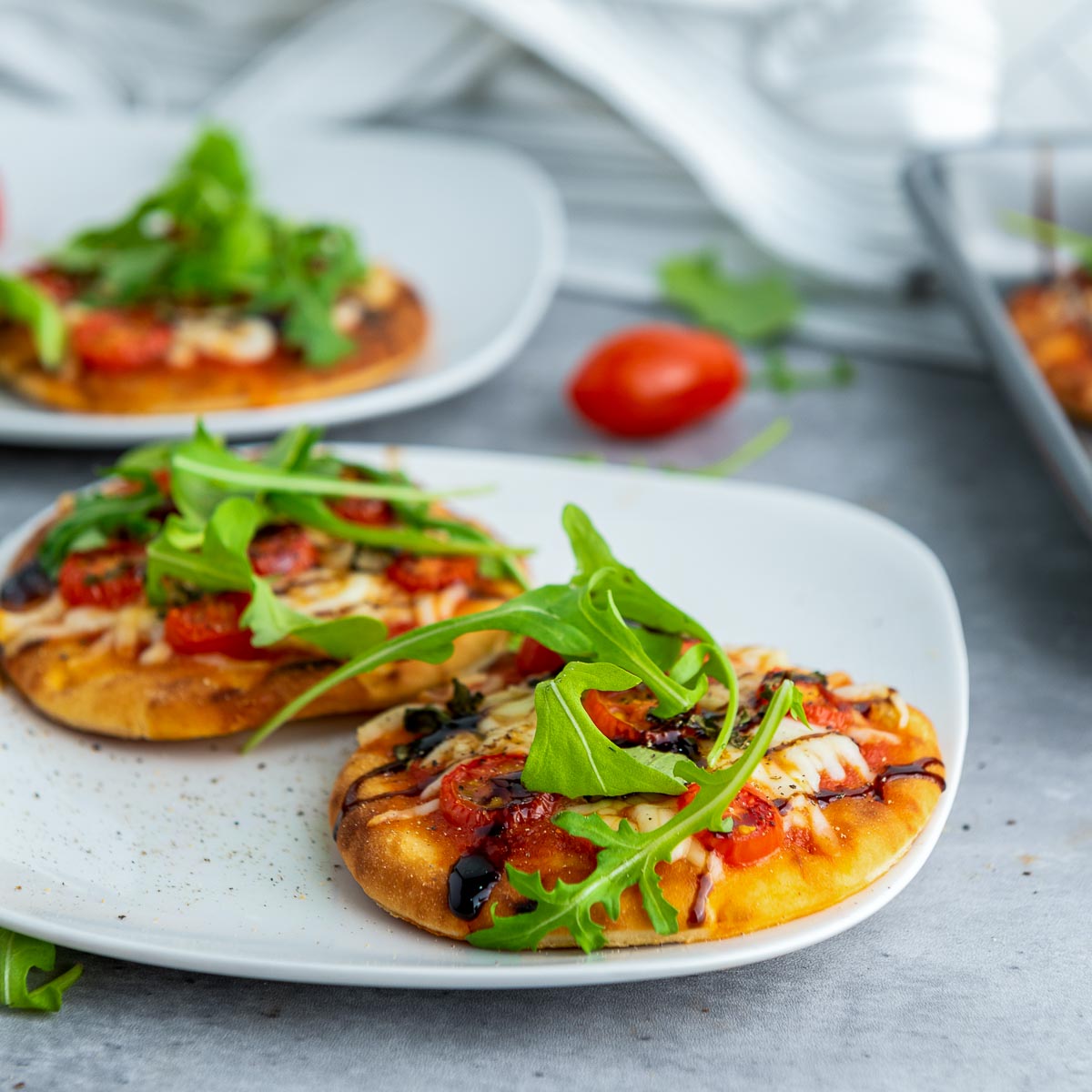 Side angle of mini Caprese pizzas served on a white plate, with arugula and tomato toppings visible.