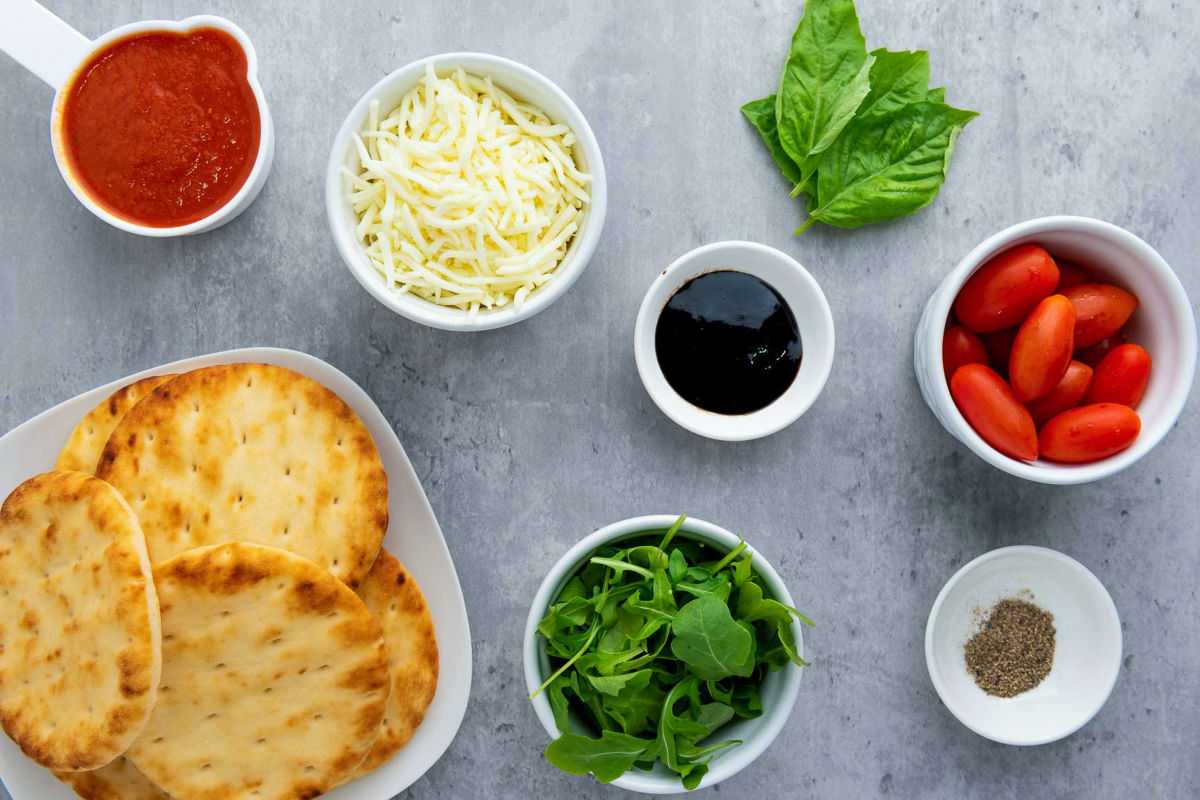 Flat lay of ingredients for mini Caprese pizza including naan bread, mozzarella, marinara sauce, tomatoes, arugula, basil leaves, balsamic glaze, and pepper.