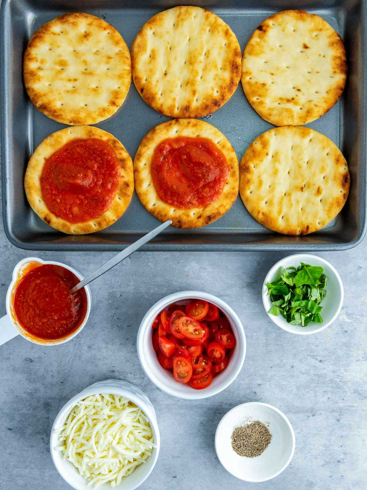 Mini flatbreads on a baking tray with two spread with tomato sauce; ingredients like shredded cheese, sliced tomatoes, and herbs are placed around.