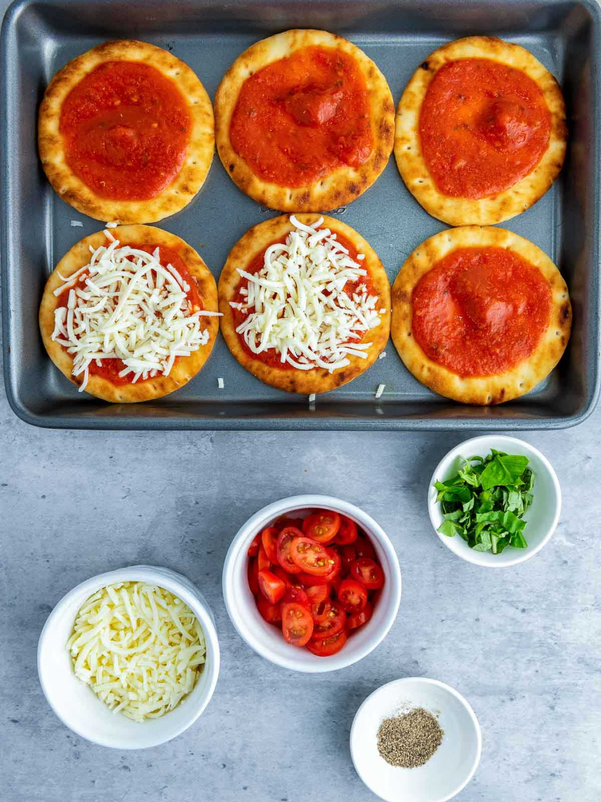Flatbreads topped with marinara and shredded mozzarella on a baking tray, with bowls of ingredients arranged below.