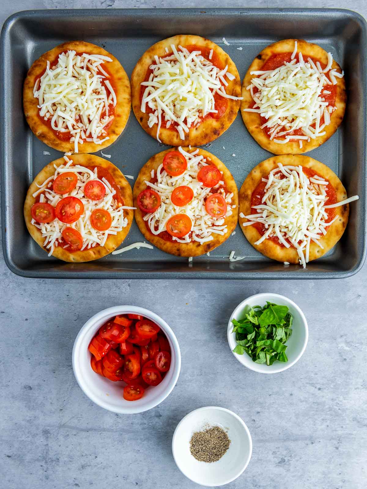 Mini pizzas topped with mozzarella and cherry tomato slices on a tray, ready for baking.