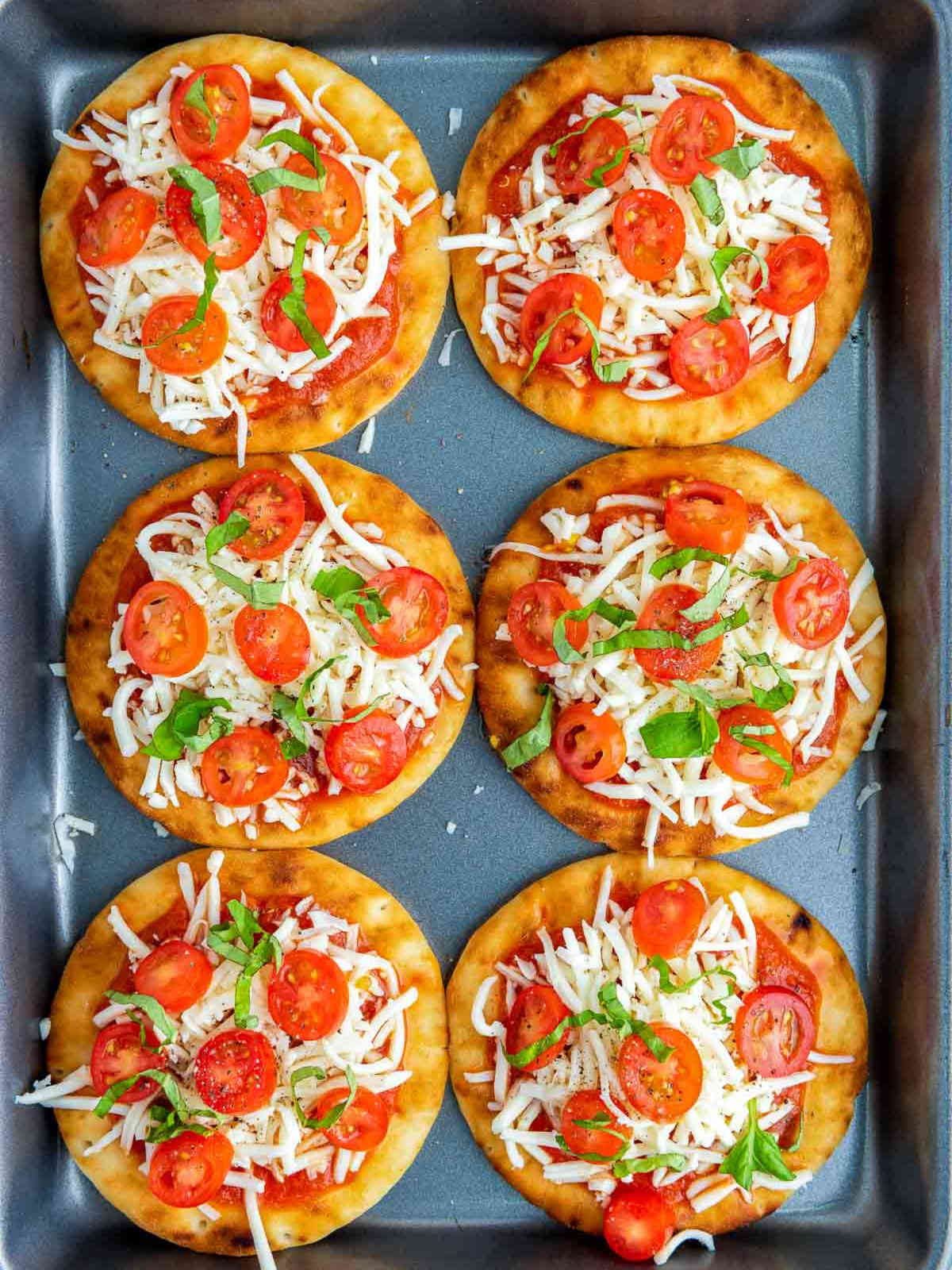 Tray of assembled pizzas topped with shredded mozzarella, tomatoes, basil, and black pepper.