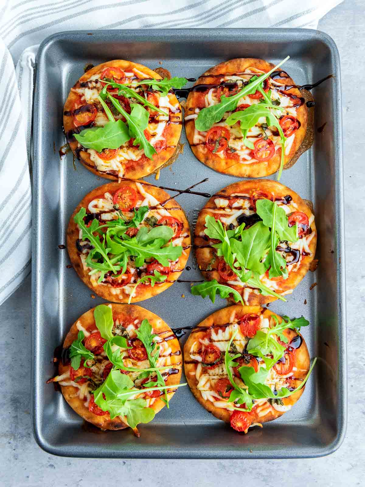 Freshly baked mini Caprese pizzas in a pan, topped with arugula and balsamic drizzle, ready to serve.