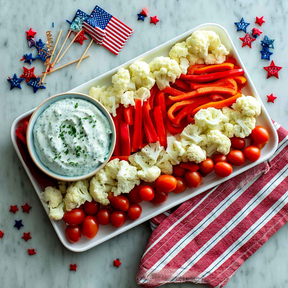 Veggie tray styled like a flag with red peppers, cauliflower, tomatoes, and ranch dip.