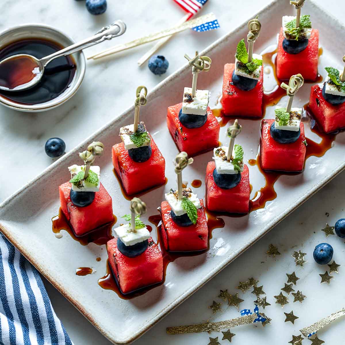 Watermelon, feta, and blueberry skewers with mint and balsamic on a white tray.