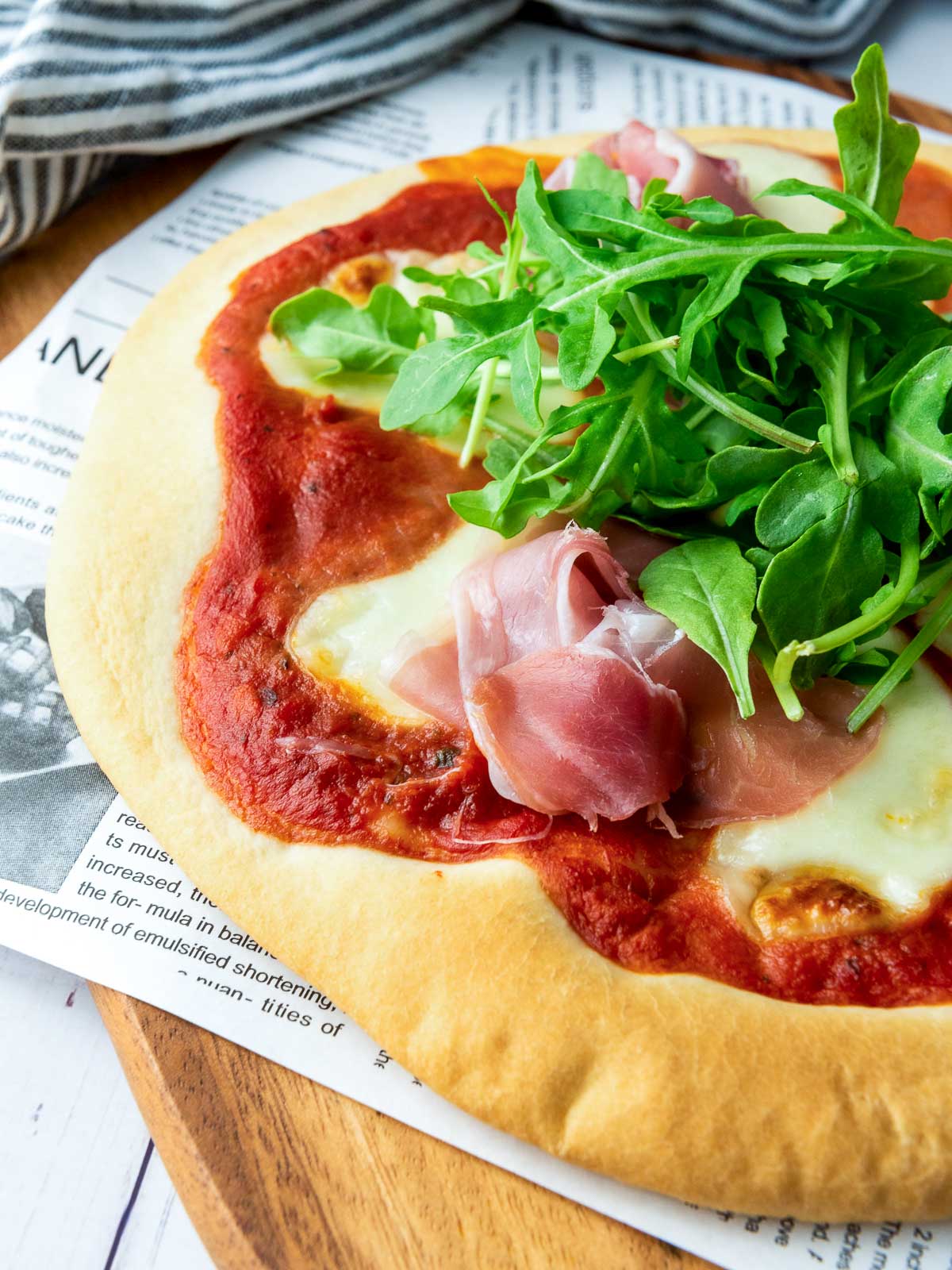 Close-up of pizza crust with melted cheese, tomato sauce, arugula, and prosciutto.