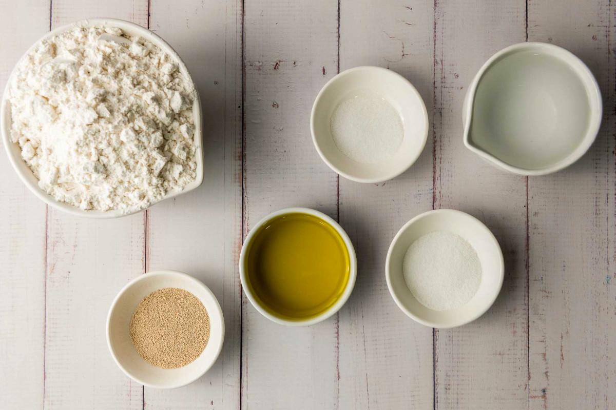 All-purpose flour, yeast, olive oil, sugar, salt, and warm water measured out in small bowls on a white wooden background.