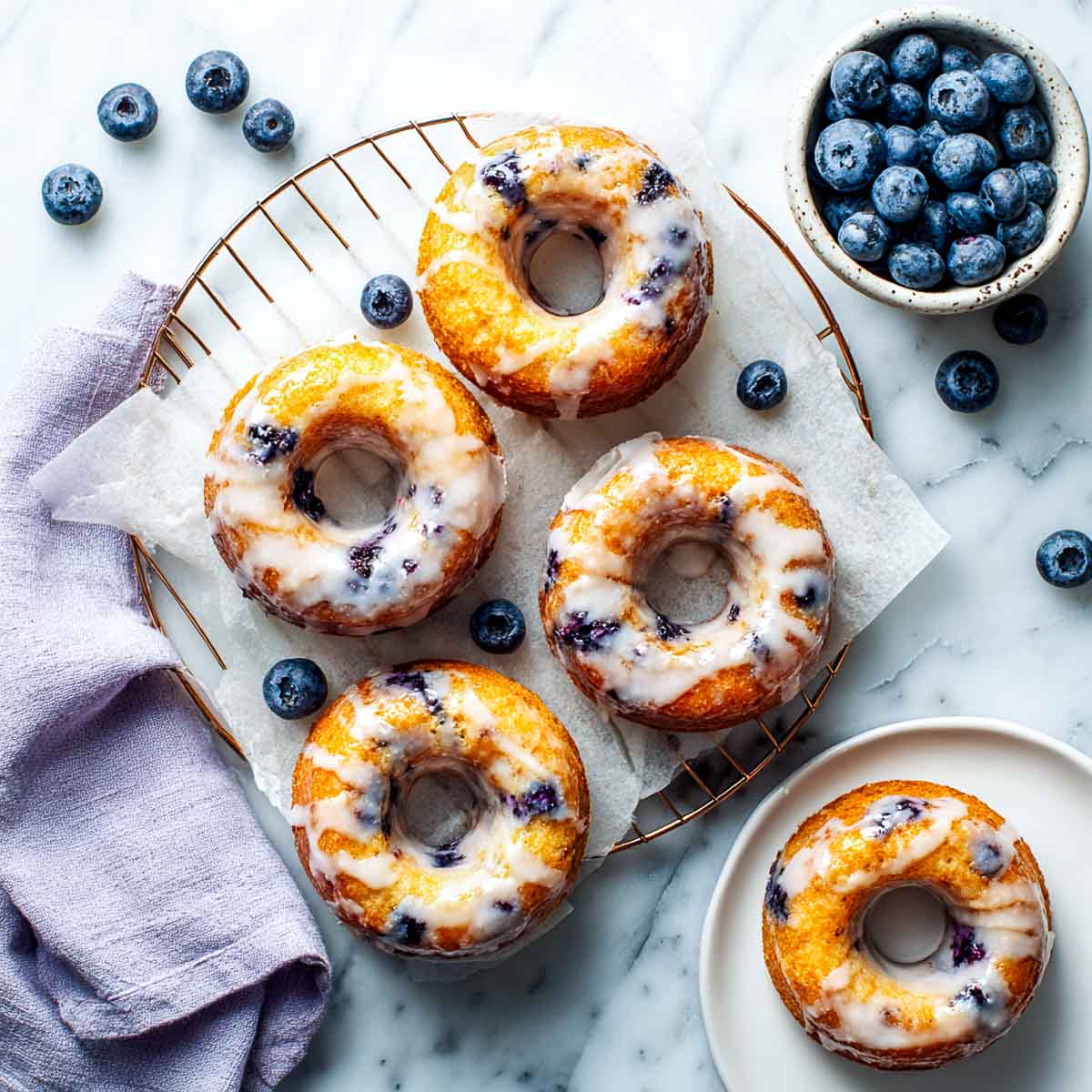 Glazed air fryer blueberry donuts cooling on a wire rack lined with parchment, surrounded by fresh blueberries.