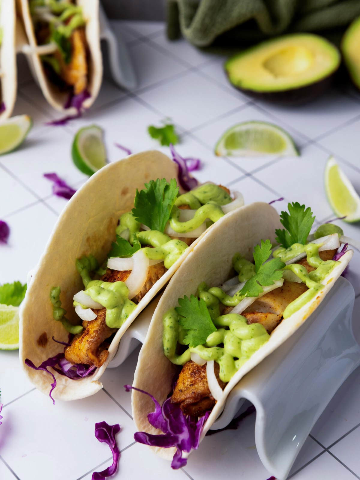 Blackened tilapia tacos on a tiled surface with lime wedges, avocados, and a green napkin in the background, topped with green sauce, onions, and cilantro.