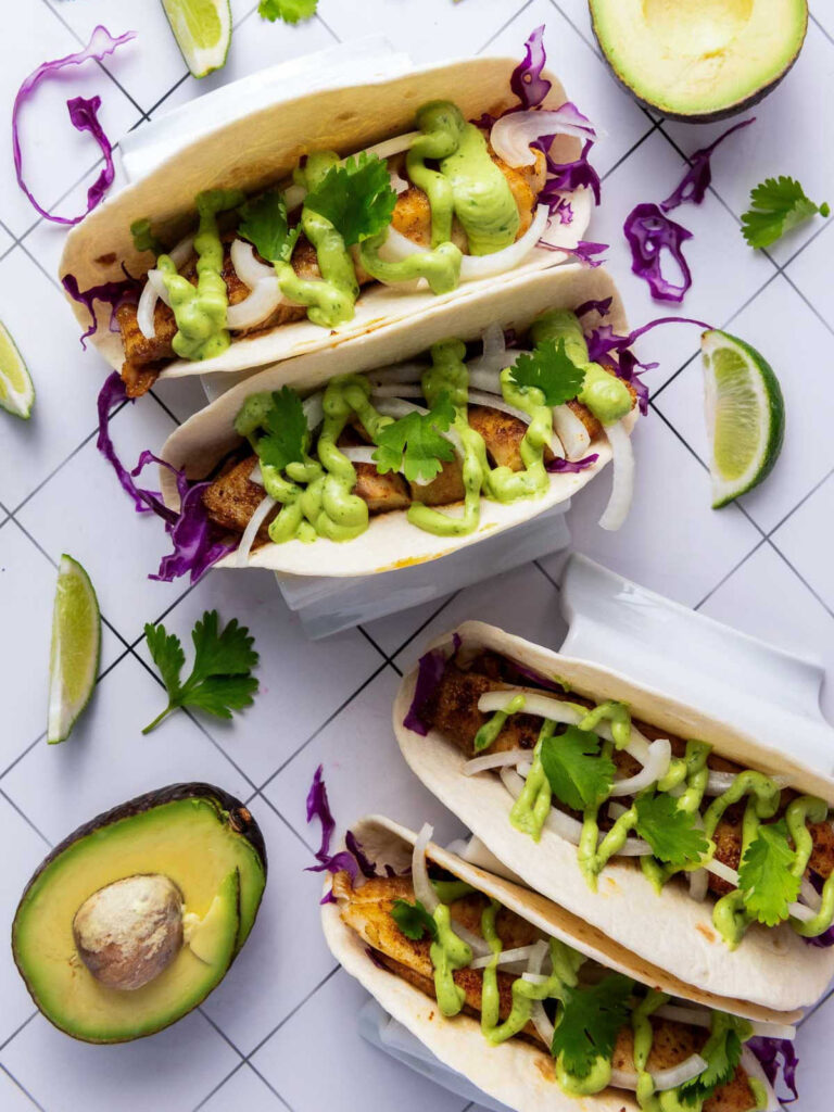 Overhead view of multiple blackened tilapia tacos filled with purple cabbage, grilled fish, onions, avocado sauce, and fresh cilantro, surrounded by limes and avocado halves.