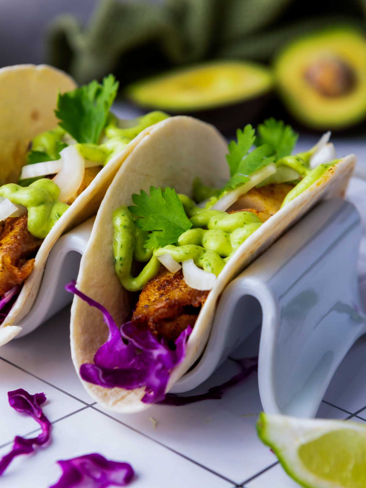 Close-up side view of blackened tilapia tacos showing crisp fish, creamy green sauce, sliced onions, and vibrant purple cabbage on a white holder.