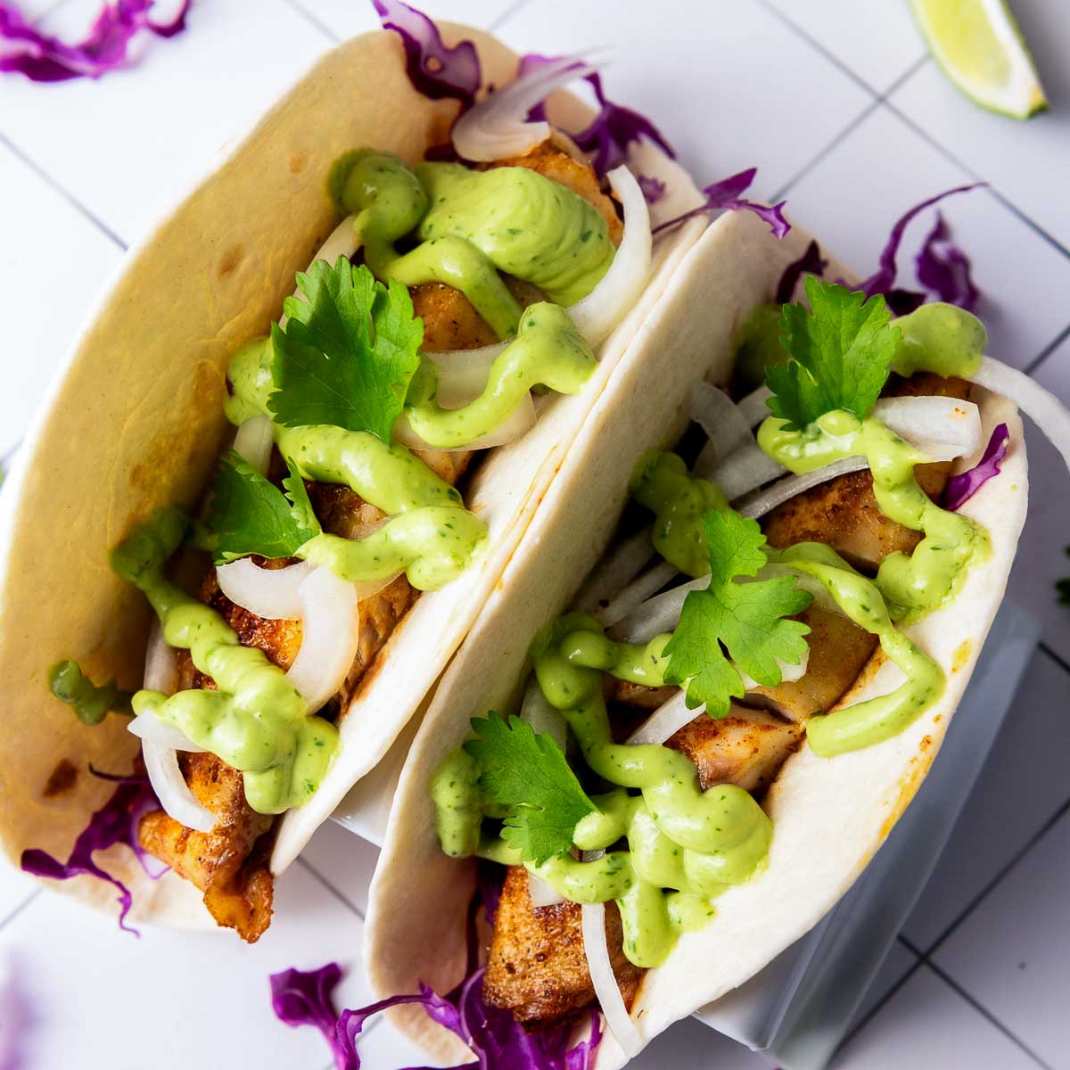 Top-down image of two blackened tilapia tacos loaded with grilled fish, onions, purple cabbage, creamy avocado sauce, and garnished with cilantro.