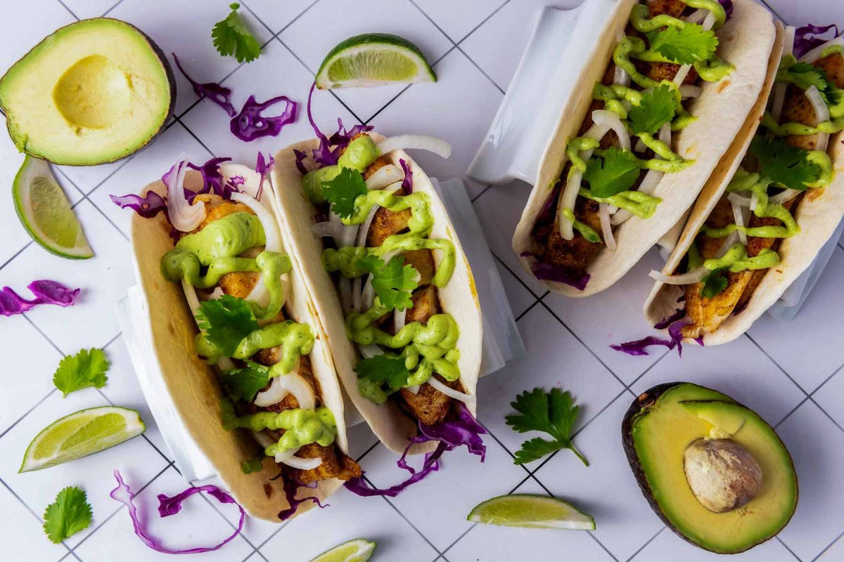 Overhead shot of assembled blackened tilapia tacos garnished with creamy green sauce, fresh cilantro, and lime wedges, styled with halved avocados and shredded cabbage.