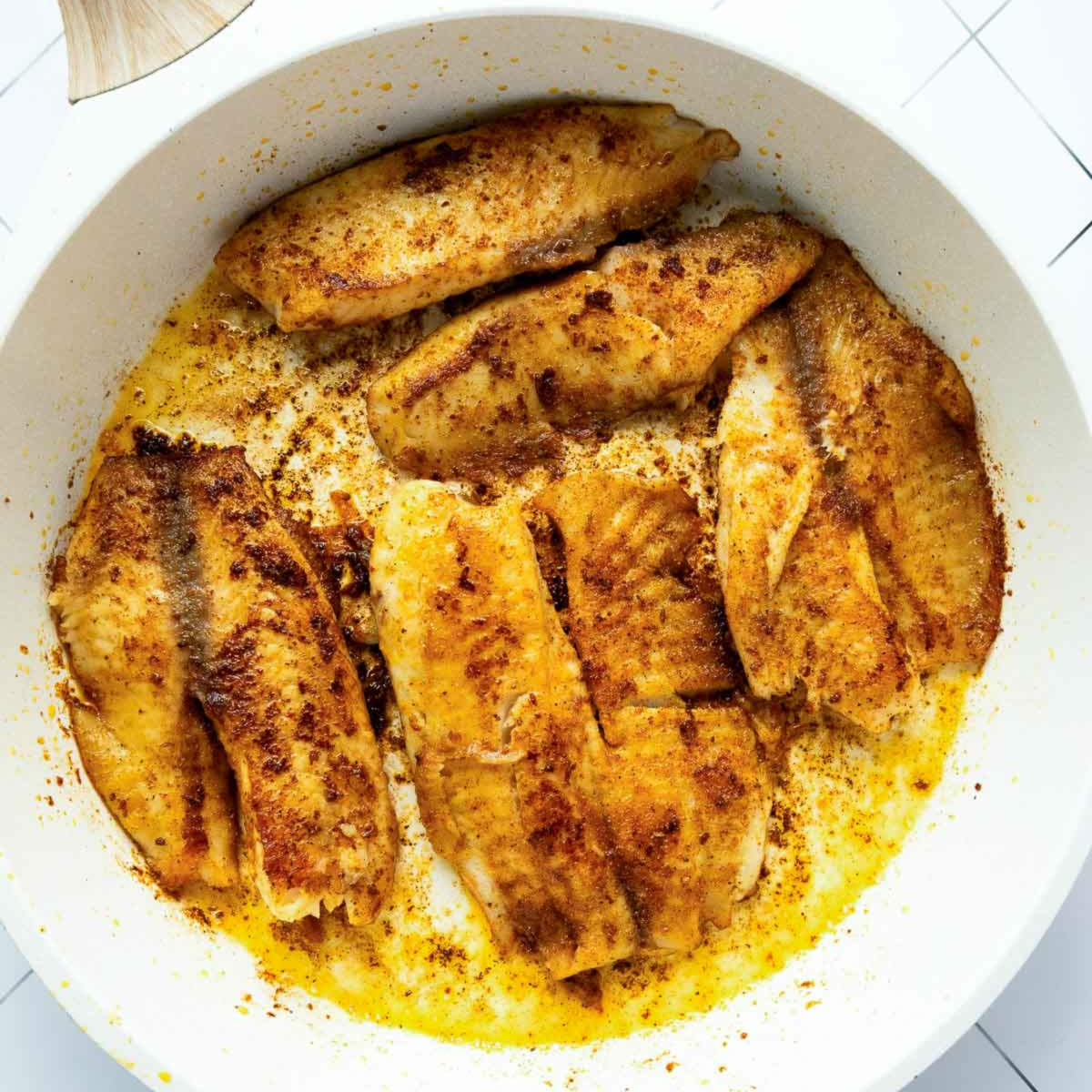 Tilapia fillets searing in a hot skillet with oil, developing a golden, seasoned crust.