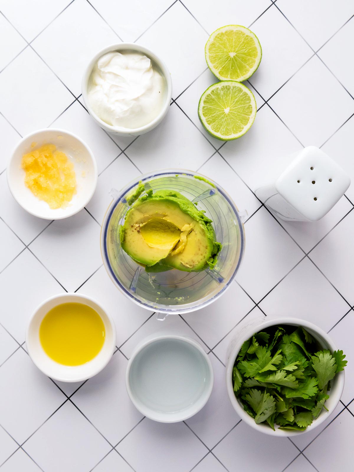 Ingredients for avocado cilantro sauce in a blender, surrounded by lime, sour cream, garlic, olive oil, water, and fresh cilantro.