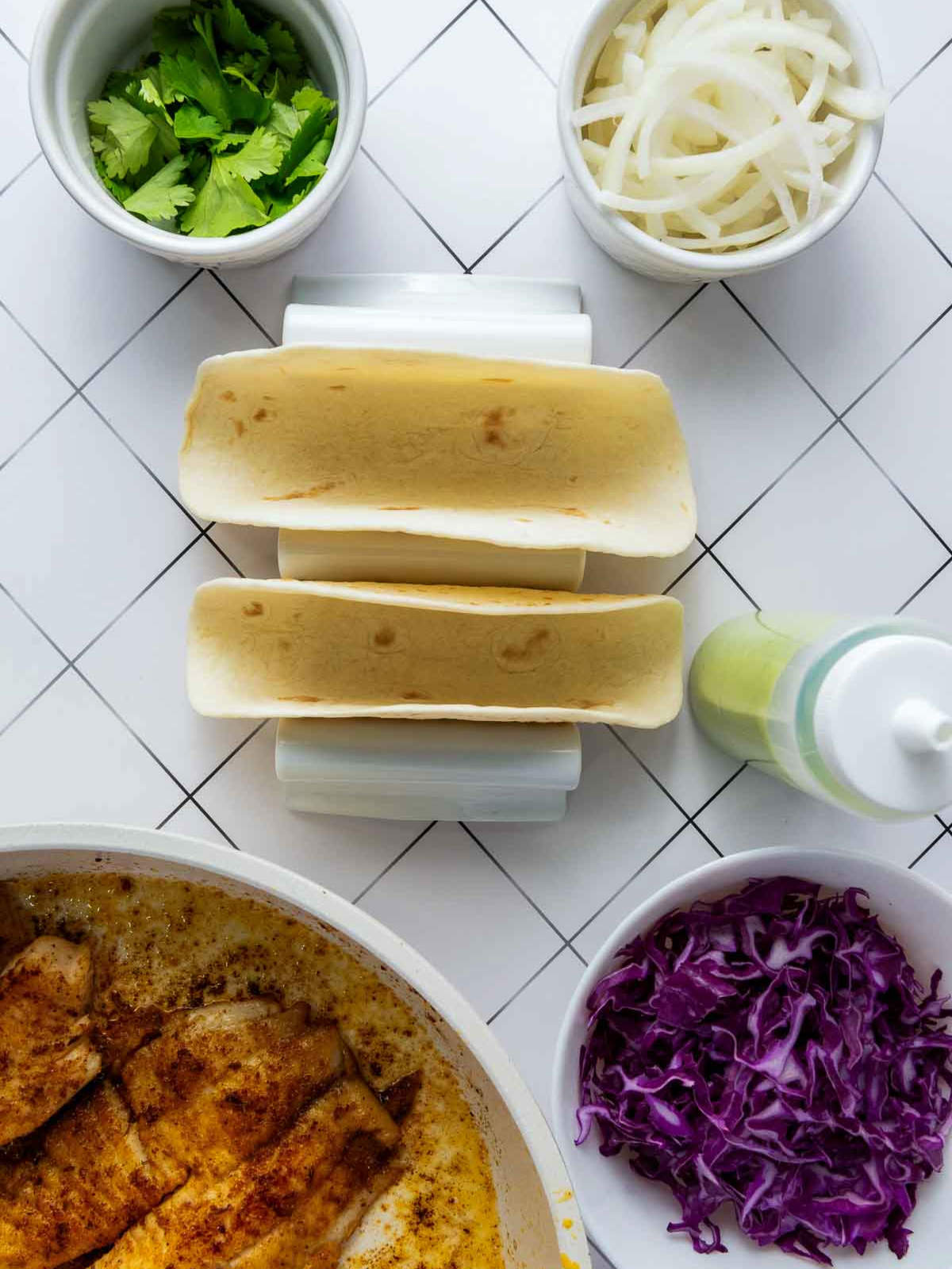 Flat tortillas set in taco holders surrounded by cooked fish, shredded cabbage, onions, cilantro, and avocado sauce bottle.